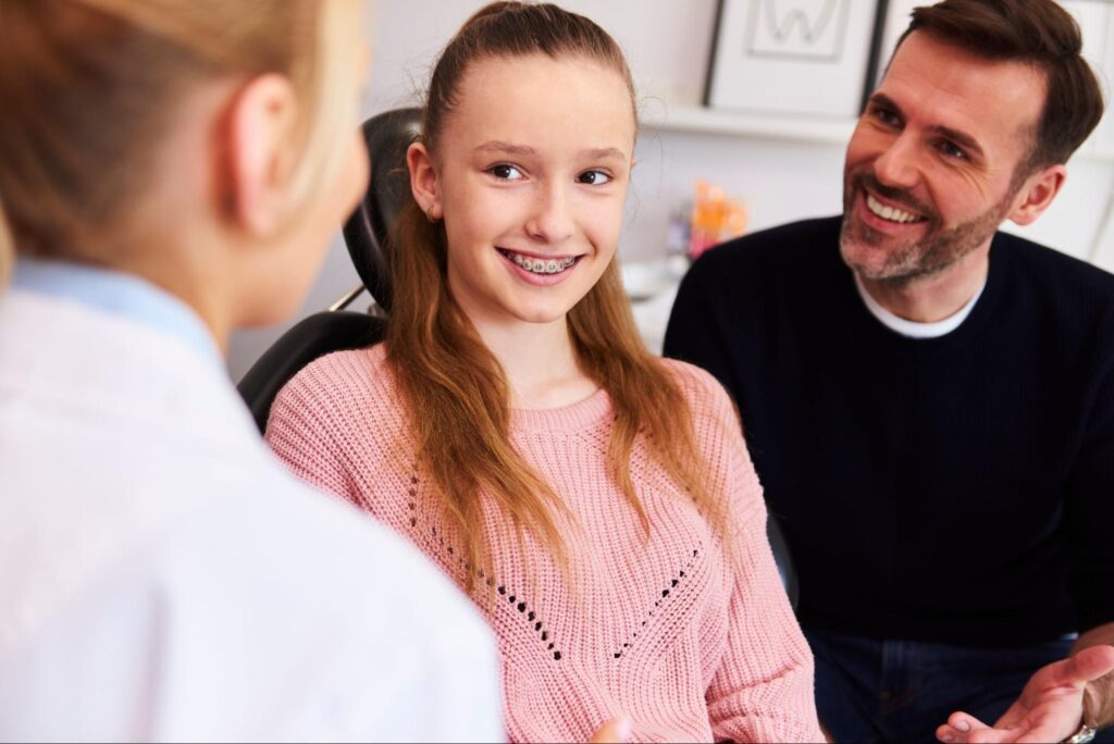 Girl with braces smiling in orthodontist chair, engaged in conversation with smiling father and orthodontist, highlighting a supportive dental environment.